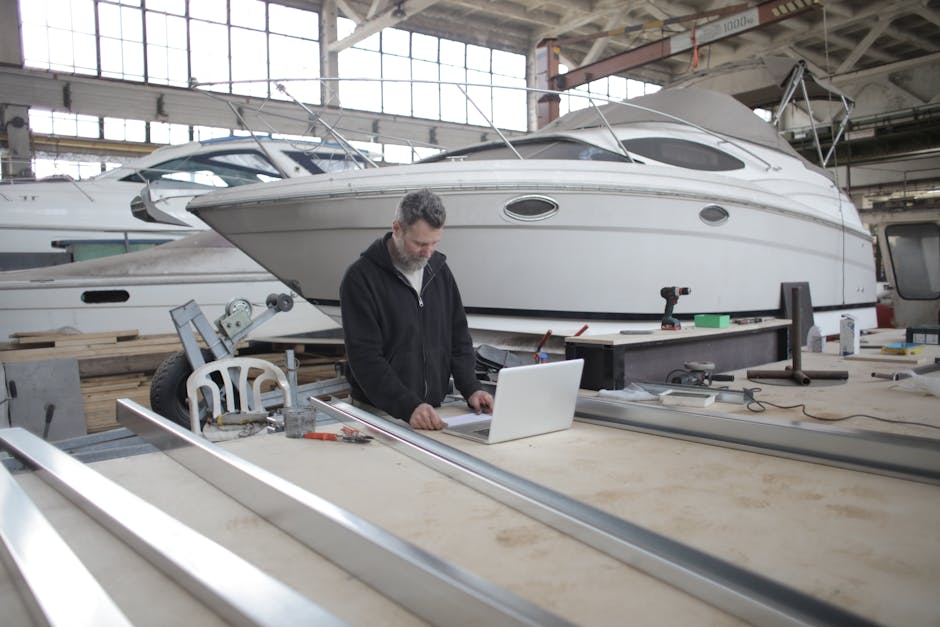 Engineer using a laptop in a boat workshop surrounded by tools and a yacht.